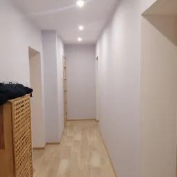 Wykończony korytarz w jasnych barwach z drewnopodobną podłogą, drewnianą szafką na buty z czarną bluzą i okularami na wierzchu oraz trzema punktowymi lampami sufitowymi.