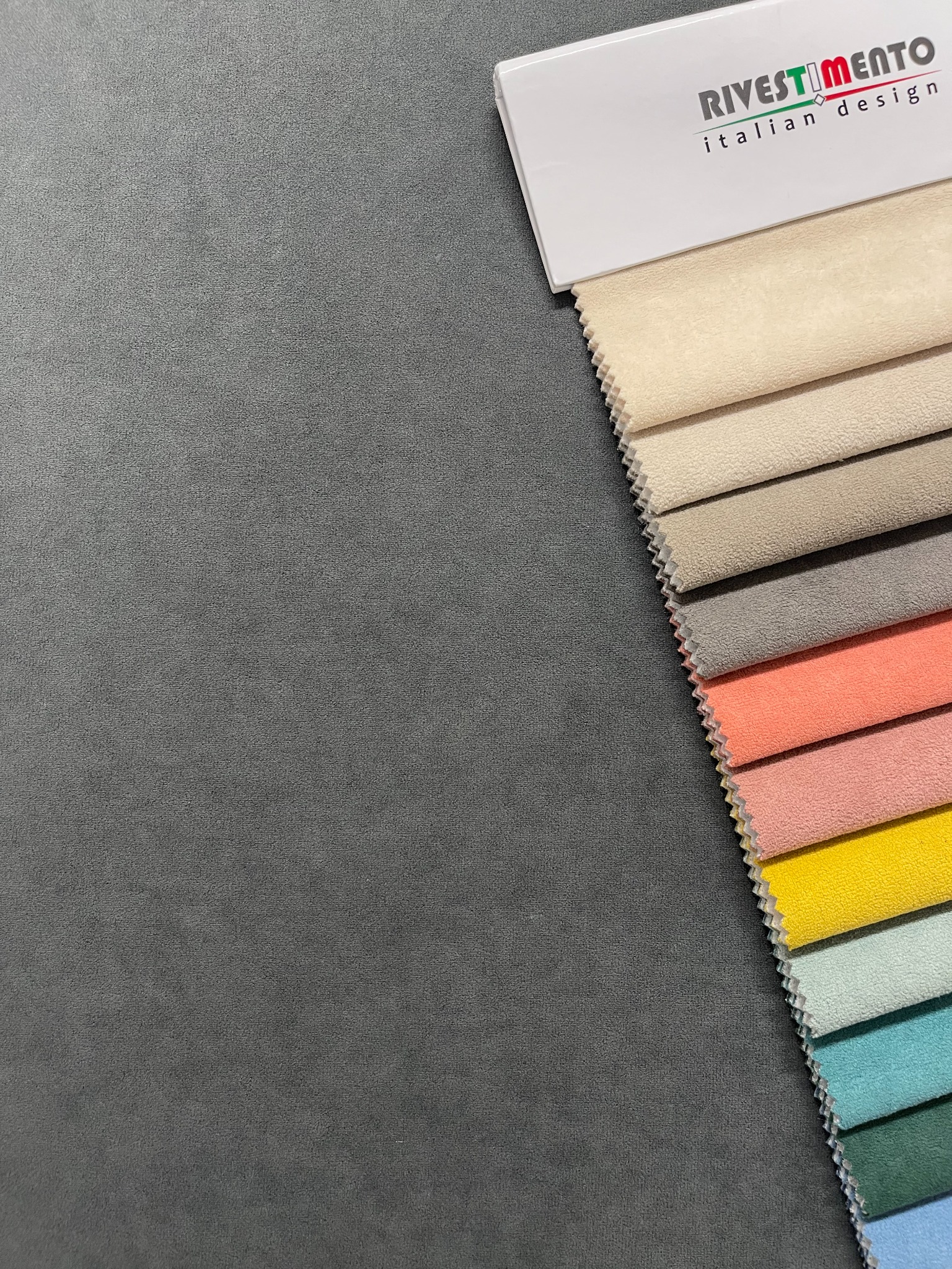 Paleta próbek tkanin w różnych kolorach, z widocznym logo 'RIVESTIMENTO italian design', ułożona na szarym tle.
