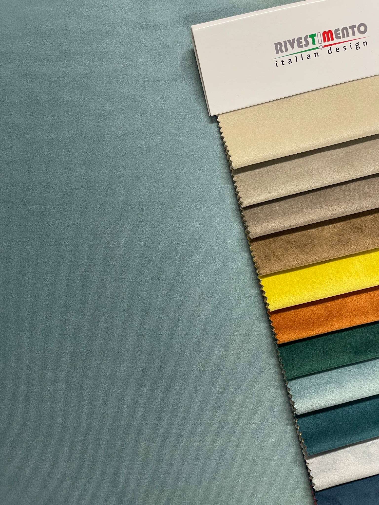 Paleta próbek tkanin w różnych kolorach i odcieniach, z widocznym logo 'RIVESTIMENTO italian design' na białym kartoniku, ułożona na tle turkusowej tkaniny.