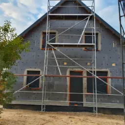 Dom w trakcie ocieplania elewacji, widoczne rusztowania, czarny styropian elewacyjny z kołkami, okna zabezpieczone folią, częściowo otynkowana podstawa budynku.
