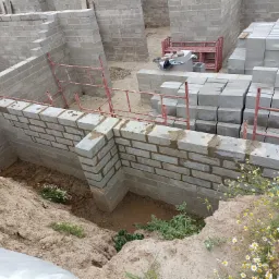 Budowa fundamentów z bloczków betonowych w Brodnicy, widoczne świeżo wymurowane ściany fundamentowe, stos bloczków oraz czerwone rusztowanie.