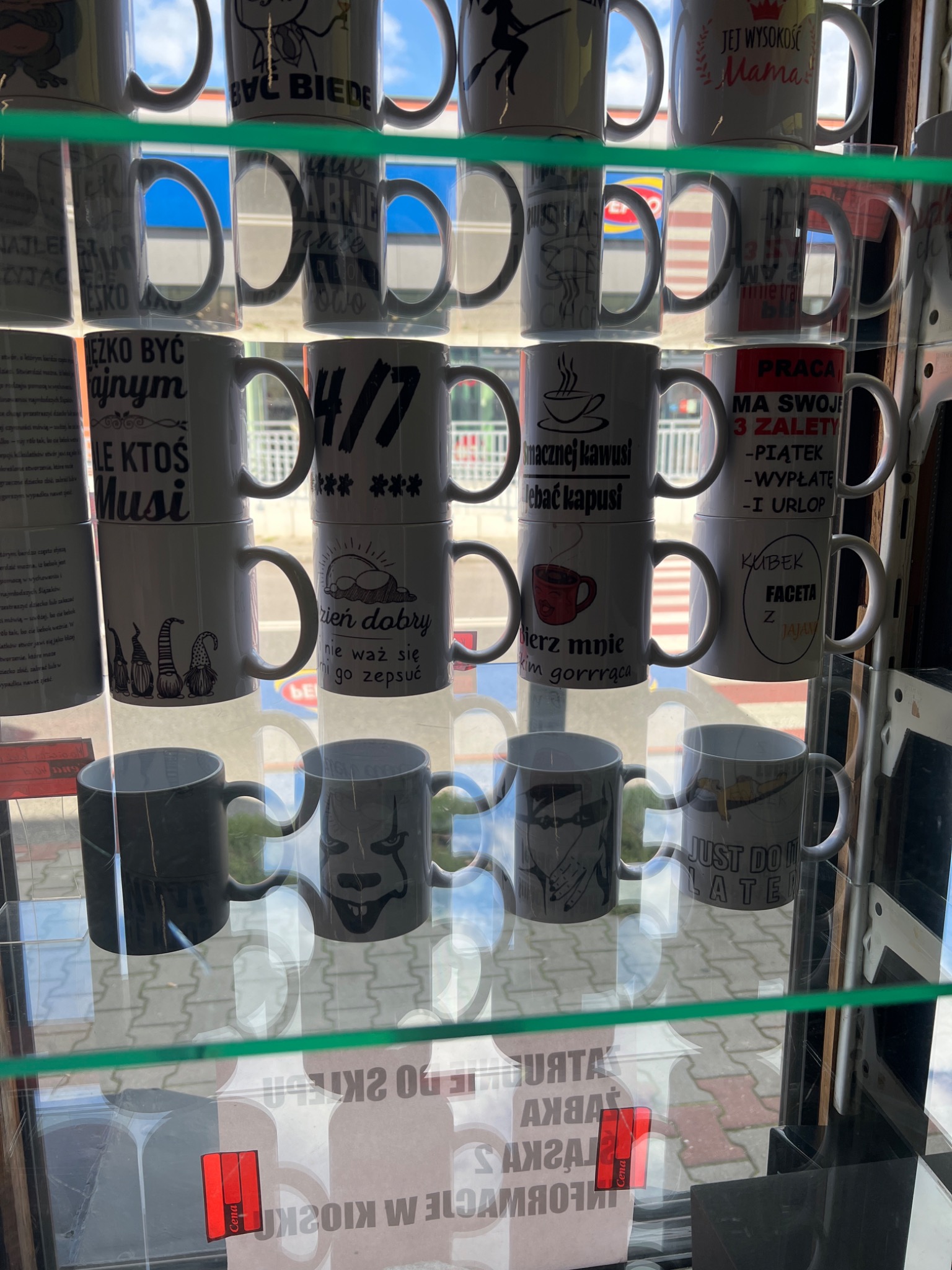 Regał z różnorodnymi kubkami z nadrukami, widoczne napisy i grafiki, w tym 'Być biedę', '4/7', 'Dzień dobry', 'Smacznej kawusi', 'Just do it later', 'Mama', 'Praca ma swoje 3 zalety', 'Kubek faceta...