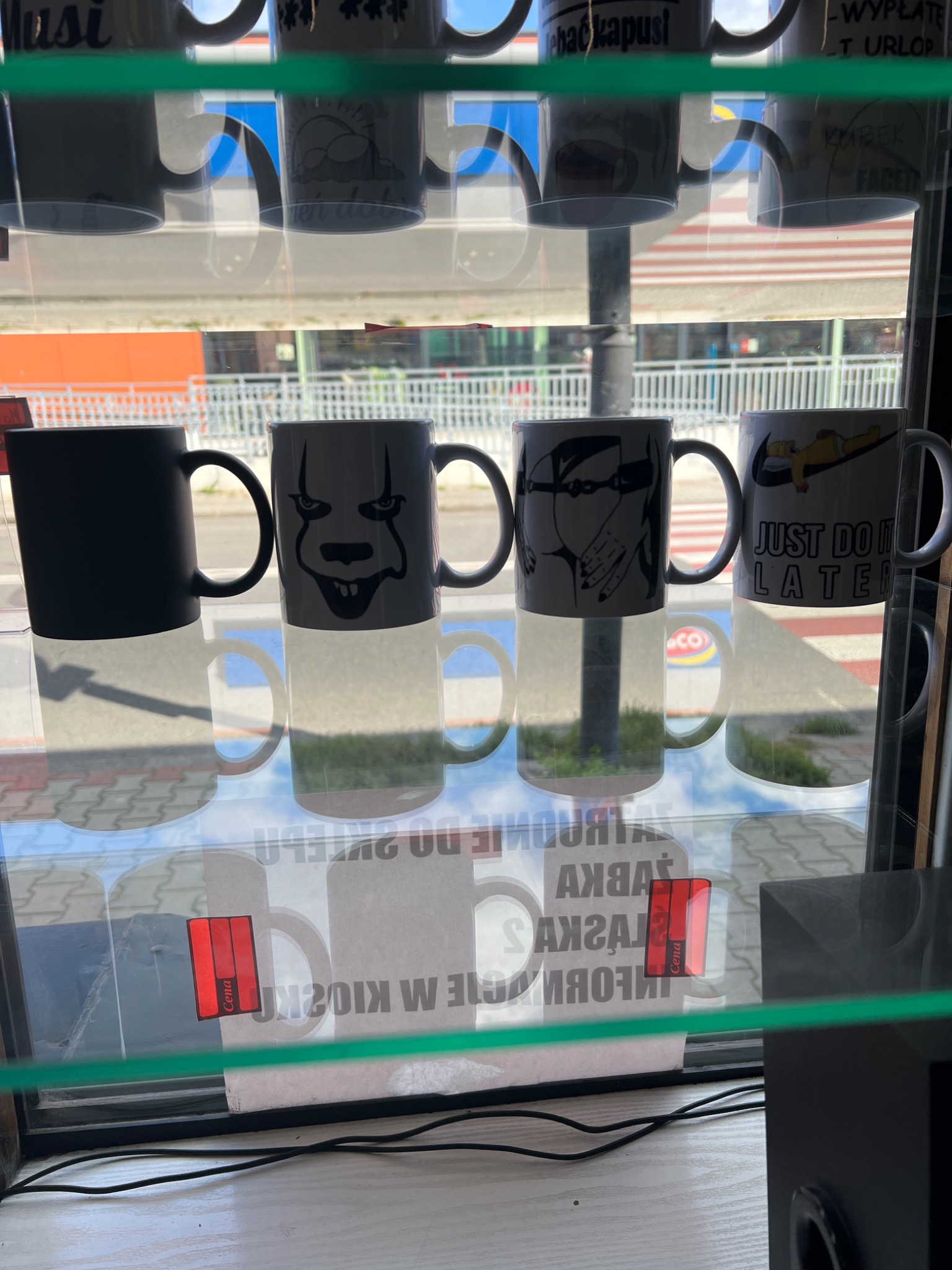 Rząd kubków z różnymi nadrukami, w tym motywem z filmu 'To' i napisem 'Just do it later', ustawionych na półce za szybą, z widocznym odbiciem ulicy.