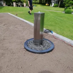 Nowoczesna fontanna ogrodowa ze stali nierdzewnej z dyszą wodną, otoczona szarymi kamieniami, na tle przygotowanego terenu pod trawnik i spacerującego mężczyzny.