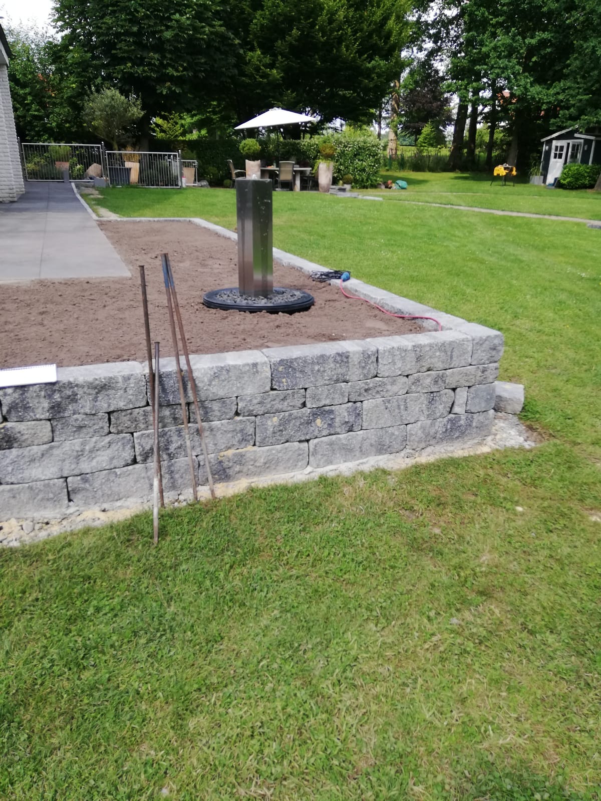 Niski murek z szarych bloczków otaczający rabatę z ziemią, w której umieszczona jest nowoczesna, metalowa fontanna; w tle trawnik, taras i meble ogrodowe.