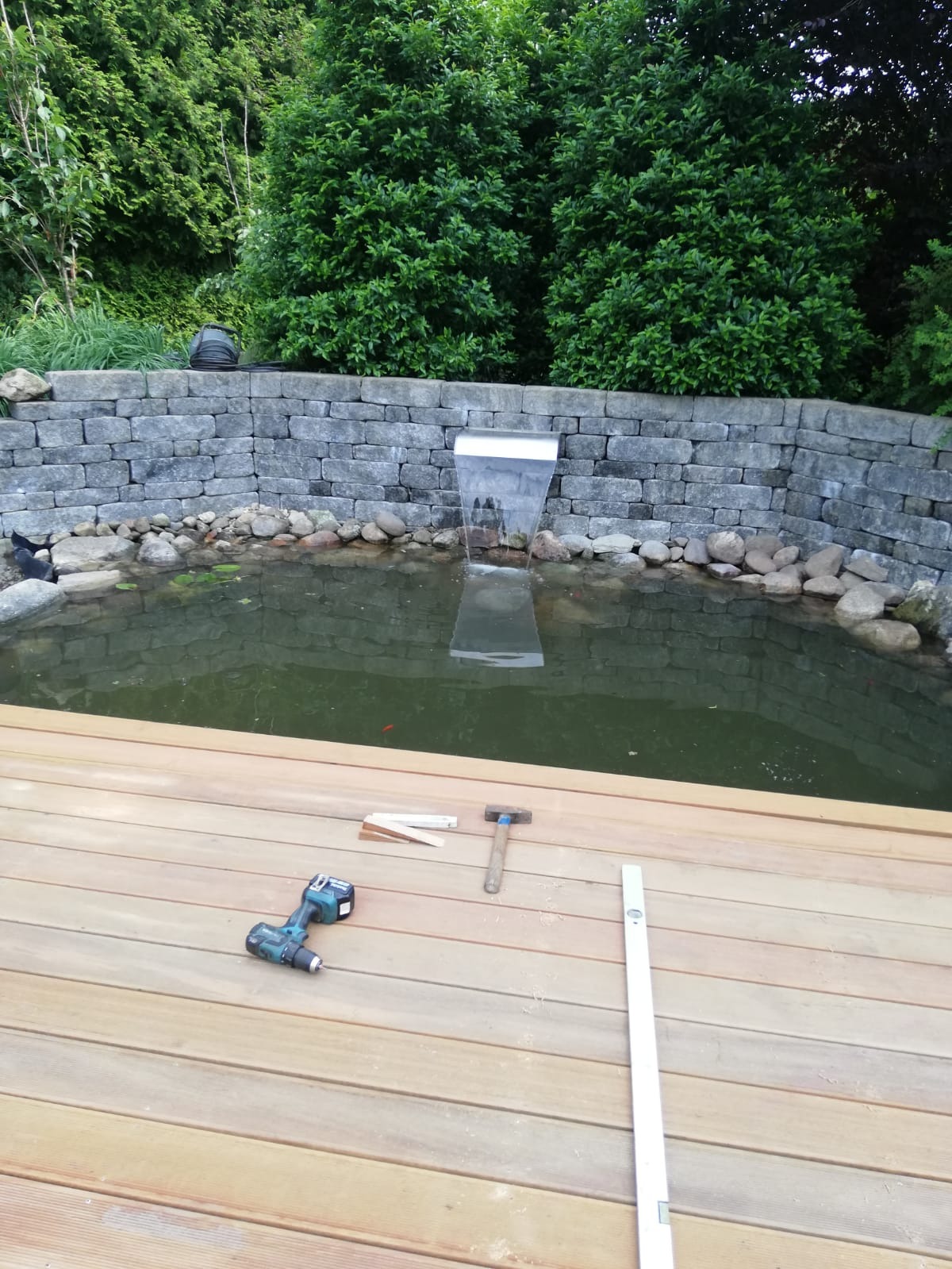 Budowa drewnianego tarasu przy oczku wodnym z wodospadem; widoczne narzędzia: wkrętarka, młotek, poziomica.