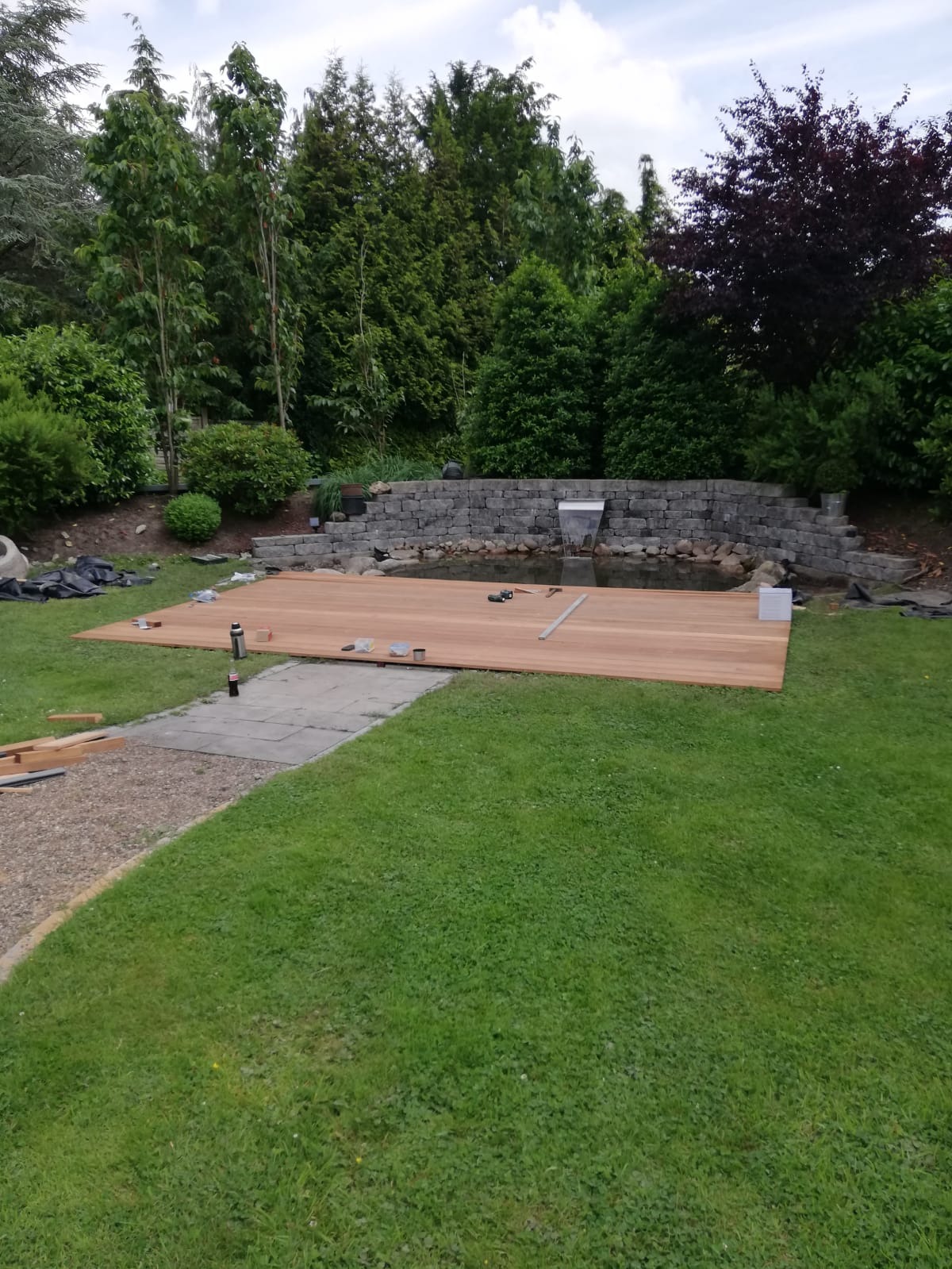 Budowa drewnianego tarasu przy oczku wodnym w ogrodzie, widoczne narzędzia i elementy konstrukcyjne na trawniku.