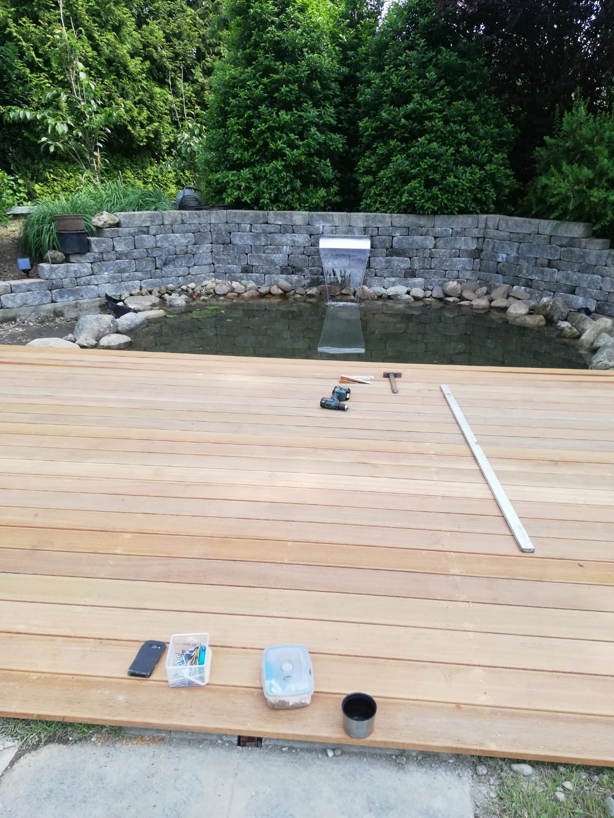 Budowa drewnianego tarasu z widokiem na oczko wodne z kaskadą, narzędzia pracy na deskach.