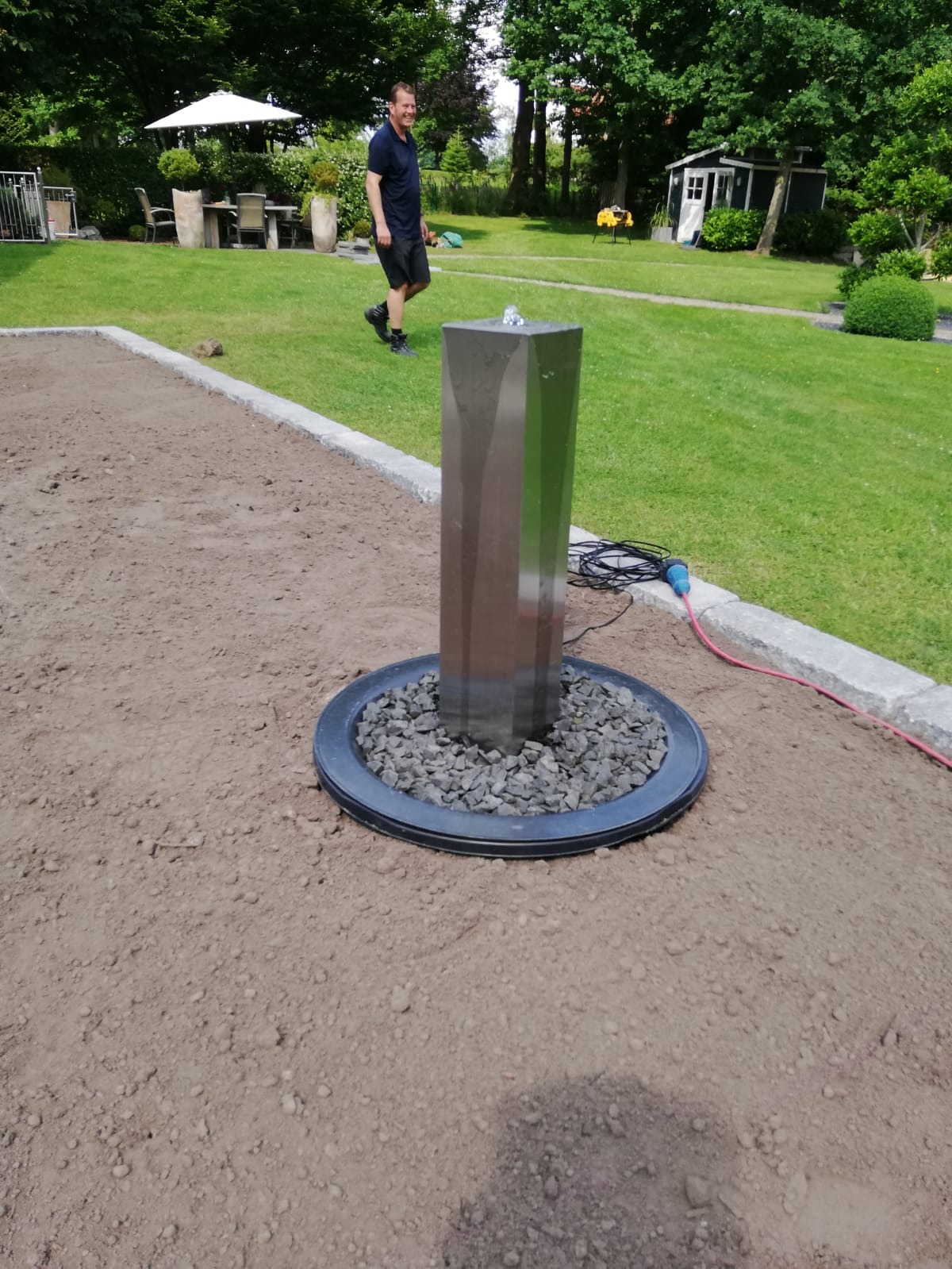 Nowoczesna fontanna ogrodowa ze stali nierdzewnej z dyszą wodną, otoczona szarymi kamieniami, na tle przygotowanego terenu pod trawnik i spacerującego mężczyzny.