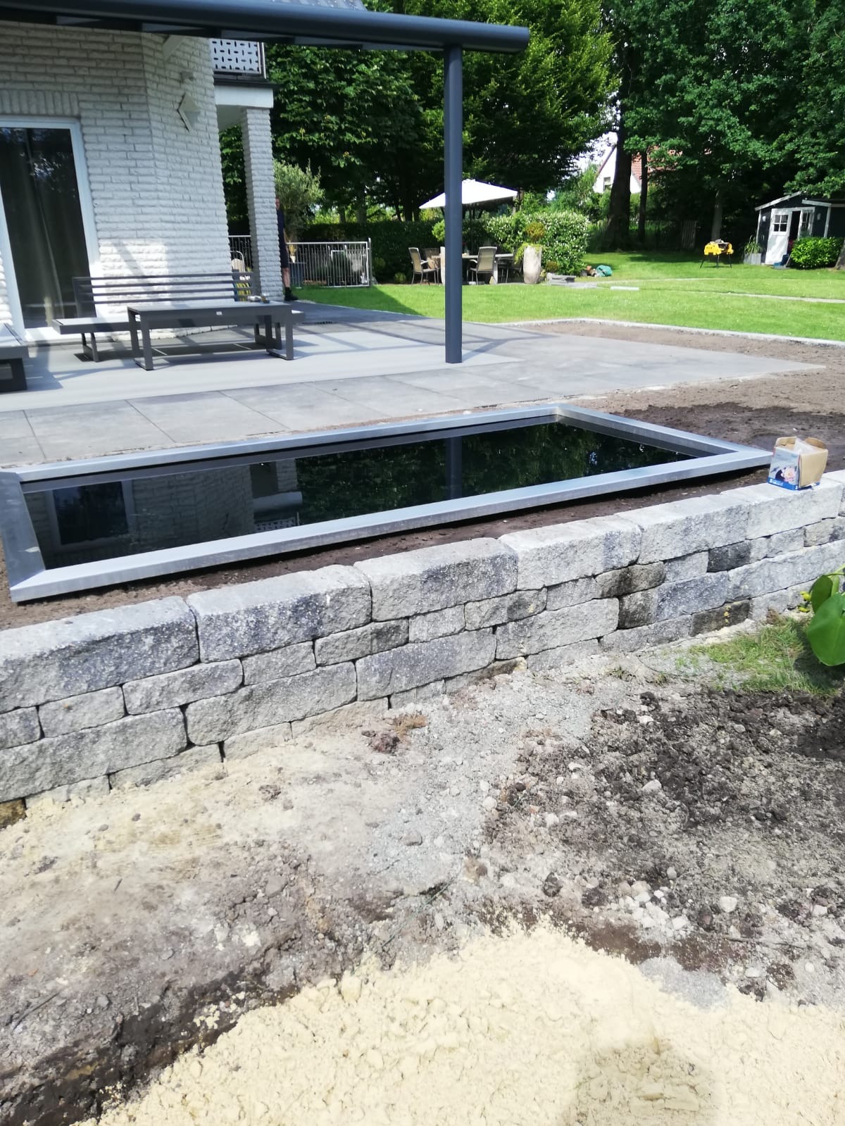 Nowoczesny zbiornik wodny z szarą ramą i kamienną obudową w ogrodzie, widoczne przygotowania do obsadzenia roślinnością, w tle taras z meblami i biały dom.