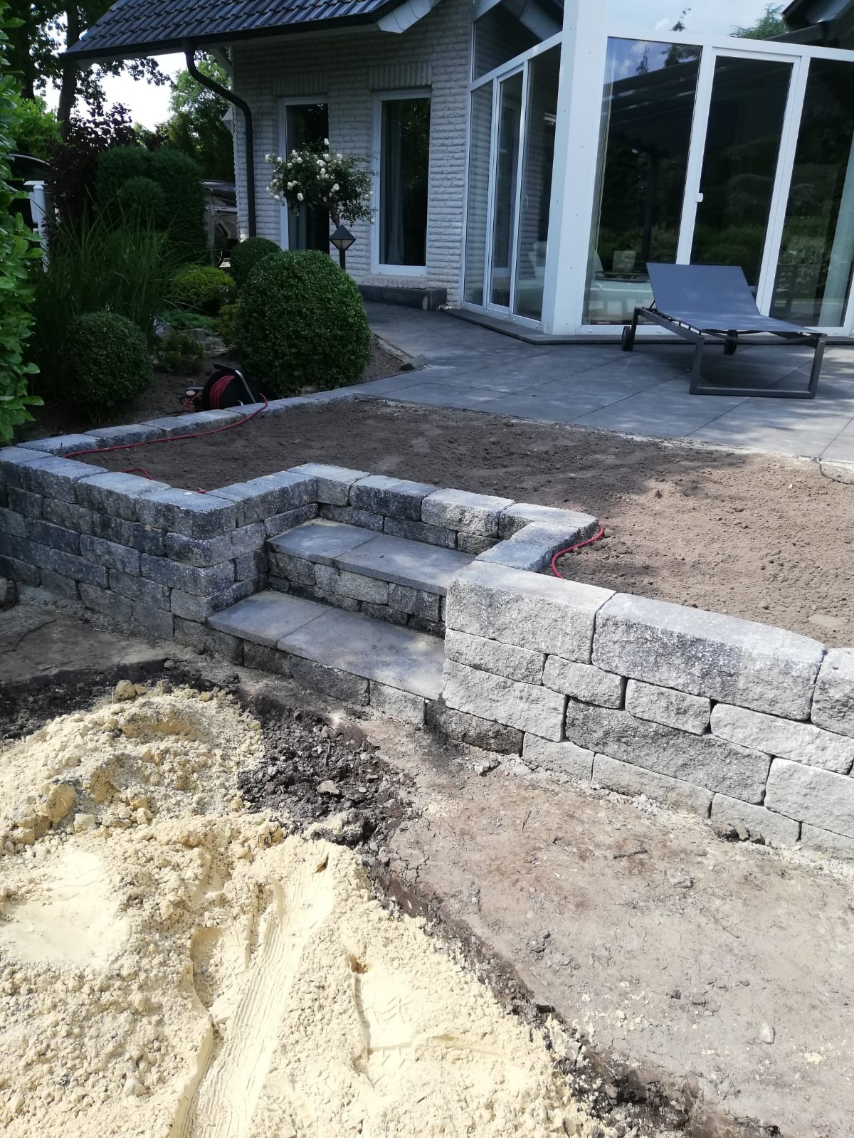 Budowa murku oporowego z kamiennymi schodami w ogrodzie, widoczne materiały budowlane i przygotowane podłoże, w tle dom z oranżerią.