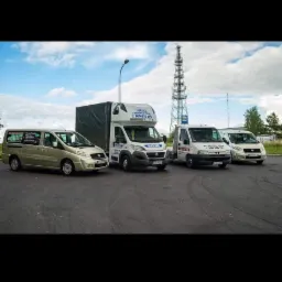 Flota czterech samochodów dostawczych zaparkowana na asfalcie, z wieżą telekomunikacyjną w tle.