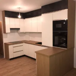 Nowoczesna kuchnia z białymi frontami szafek, drewnianymi blatami i zabudową piekarnika oraz mikrofalówki, uzupełniona o czarne wykończenia górnych szafek i sprzętu AGD.