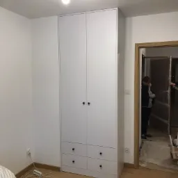 Biała szafa z czterema drzwiami i czterema szufladami z okrągłymi, czarnymi uchwytami, stojąca w pokoju z drewnianą podłogą; w tle widoczne drzwi do innego pomieszczenia i osoba.