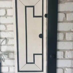 Rewizja wodomierzowa z kremową płytką w geometryczny wzór, wkomponowana w białą ścianę z cegły, z czarną ramą i widocznym elementem mocującym.