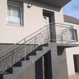 Dwukondygnacyjny budynek z elewacją w odcieniach beżu i brązu, z zewnętrznymi schodami pokrytymi szarym lastryko i metalową balustradą.