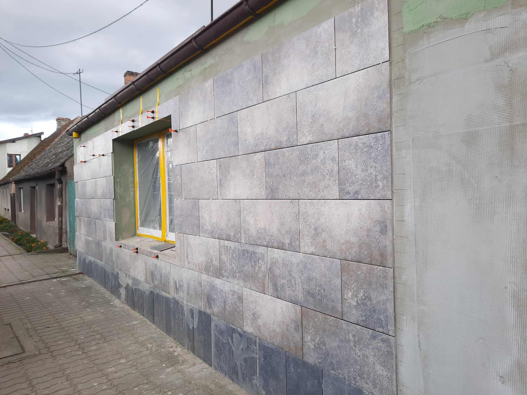 Montaż płytek elewacyjnych imitujących kamień na ścianie budynku z oknem zabezpieczonym folią, widoczne elementy mocujące i fragment przygotowanej powierzchni pod elewację.