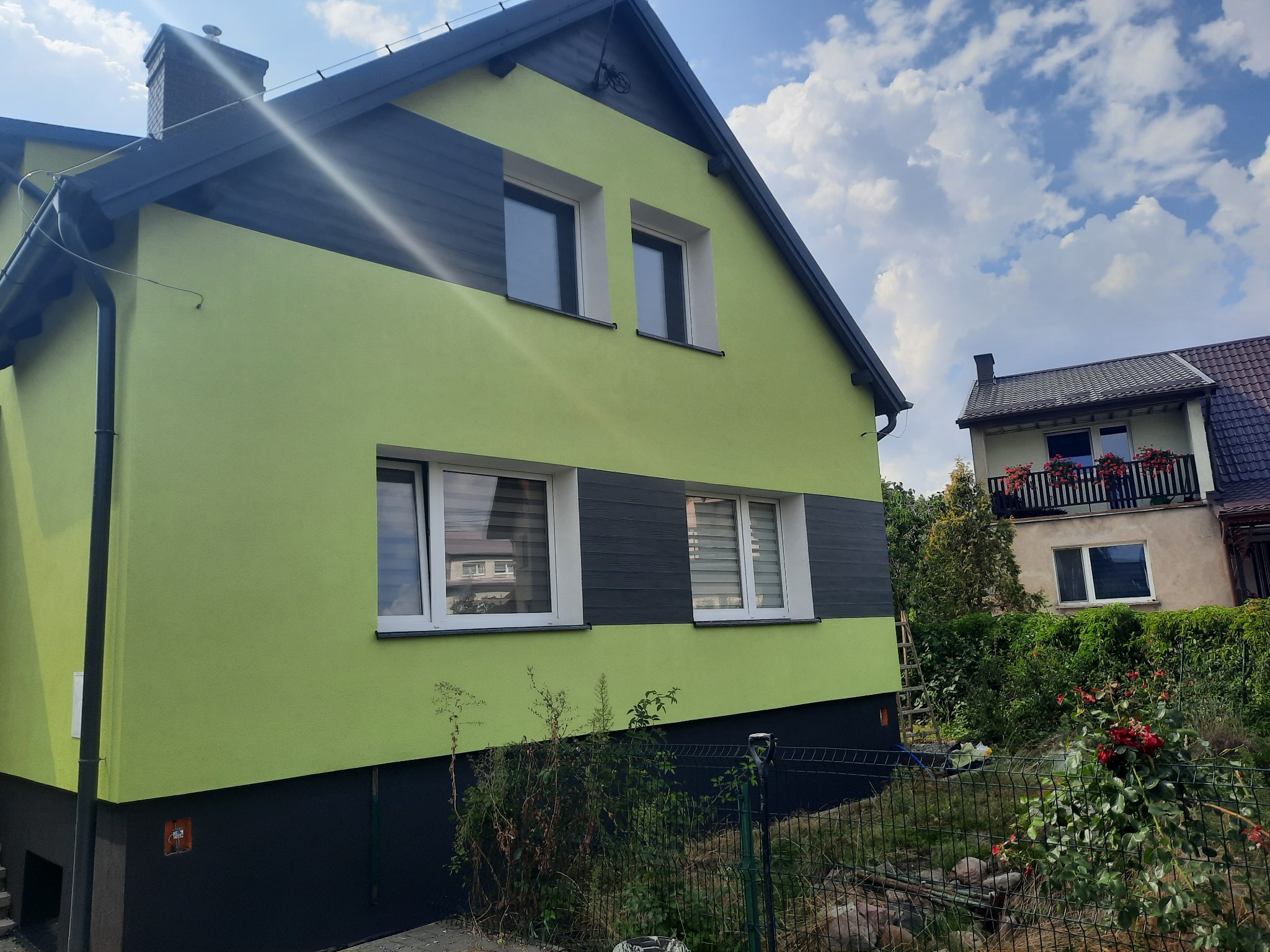 Dom z jasnozieloną elewacją i ciemnoszarymi akcentami, widok z boku na tle błękitnego nieba z chmurami.