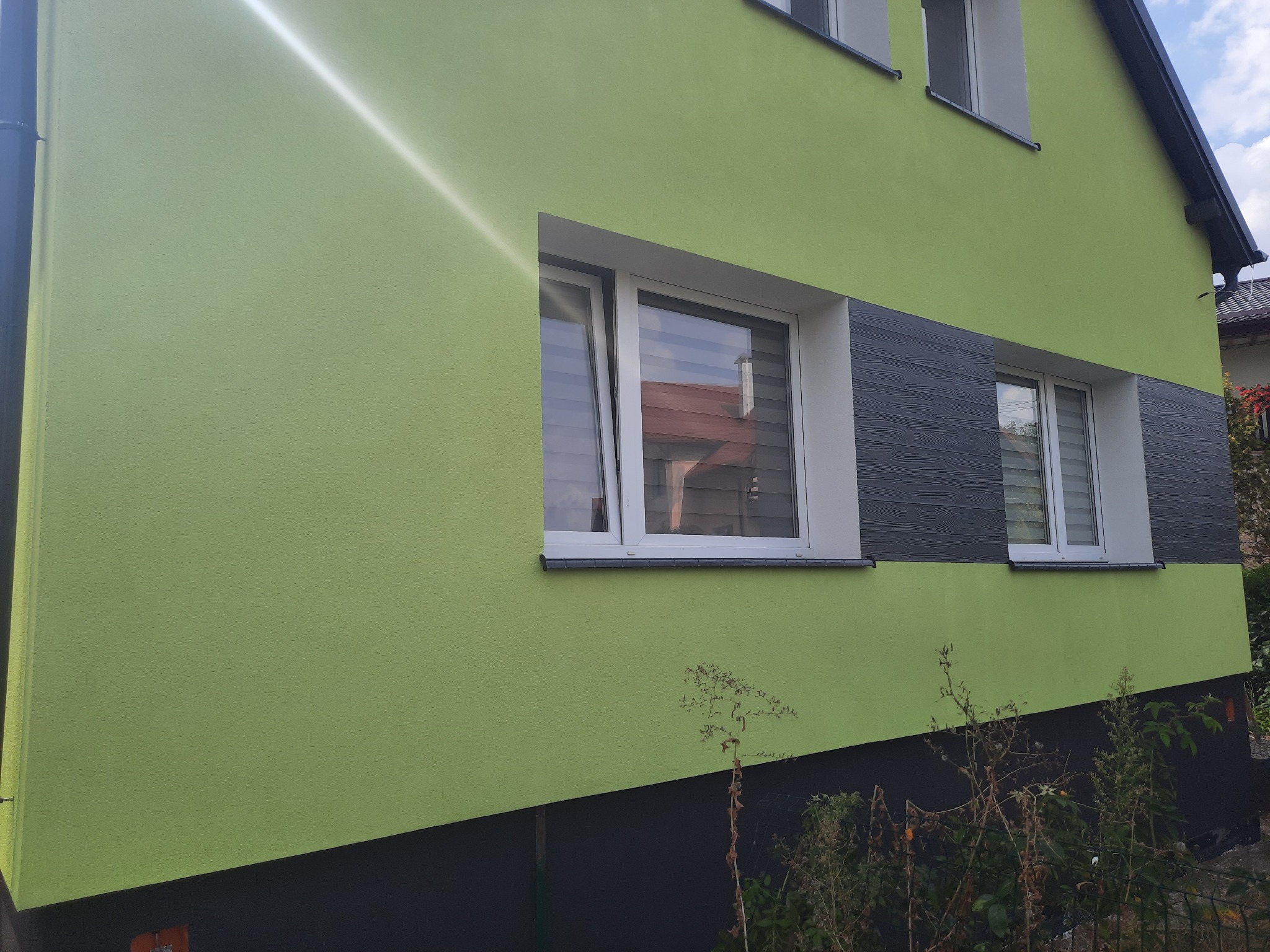 Dom z elewacją w odcieniach zieleni i szarości, z widocznymi oknami z roletami, zieleń przed budynkiem.