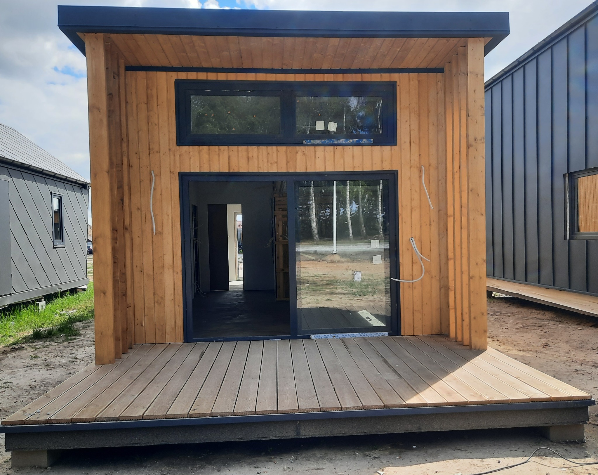 Surowa drewniana elewacja modułowego domu z czarnymi ramami okiennymi i przesuwnymi drzwiami, widok na wnętrze w trakcie wykańczania i drewniany taras.
