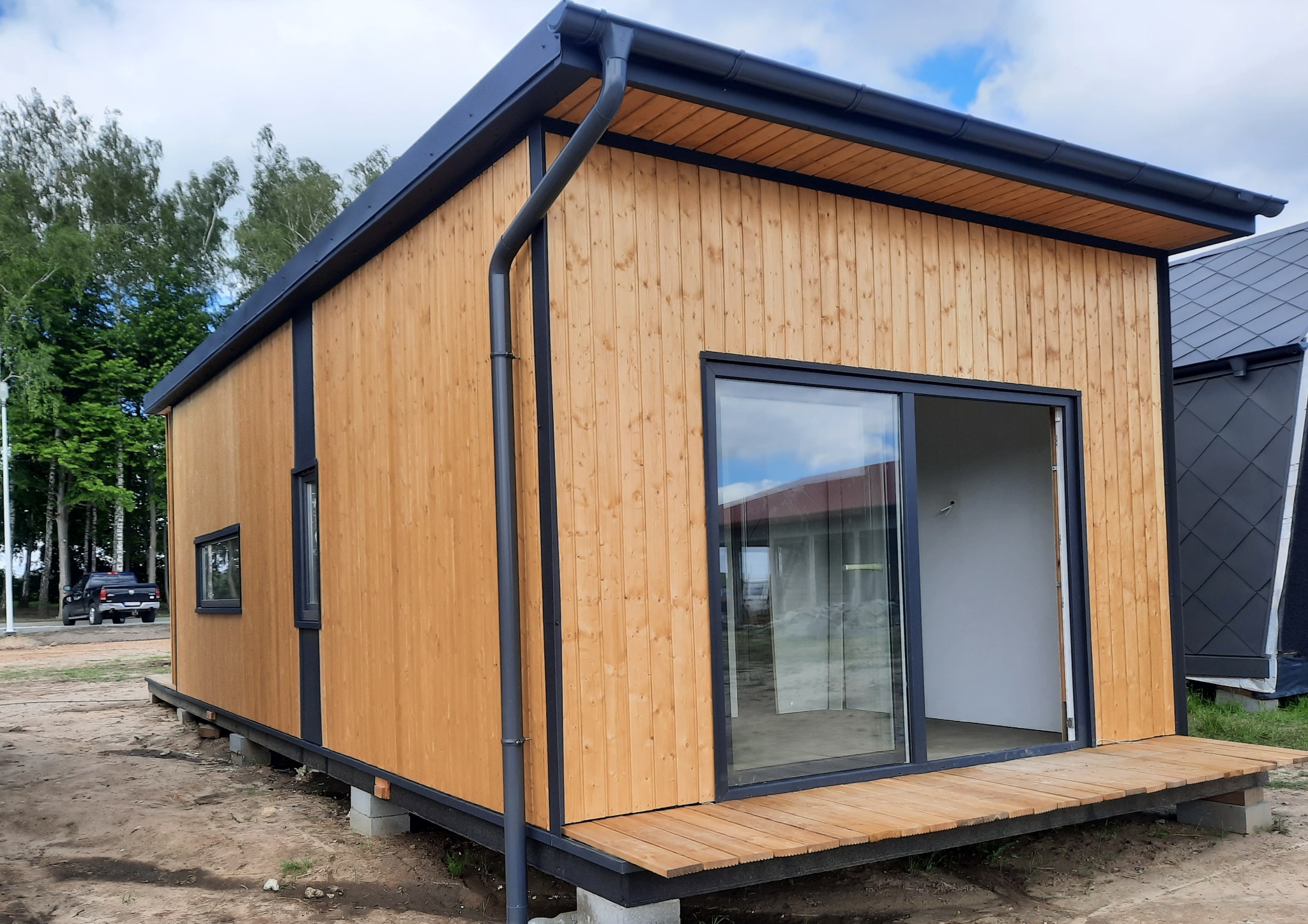 Gotowy, drewniany domek modułowy z czarnymi ramami okiennymi i orynnowaniem, posadowiony na betonowych bloczkach.