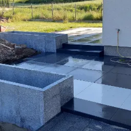 Niwelacja terenu i układanie ciemnej kostki brukowej w Krakowie, widoczne betonowe koryta i koparka na tle zielonego ogrodu.