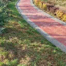 Czerwona kostka brukowa tworząca ścieżkę w ogrodzie, otoczona zielenią i prowadząca do bramy z dwoma osobami w oddali.