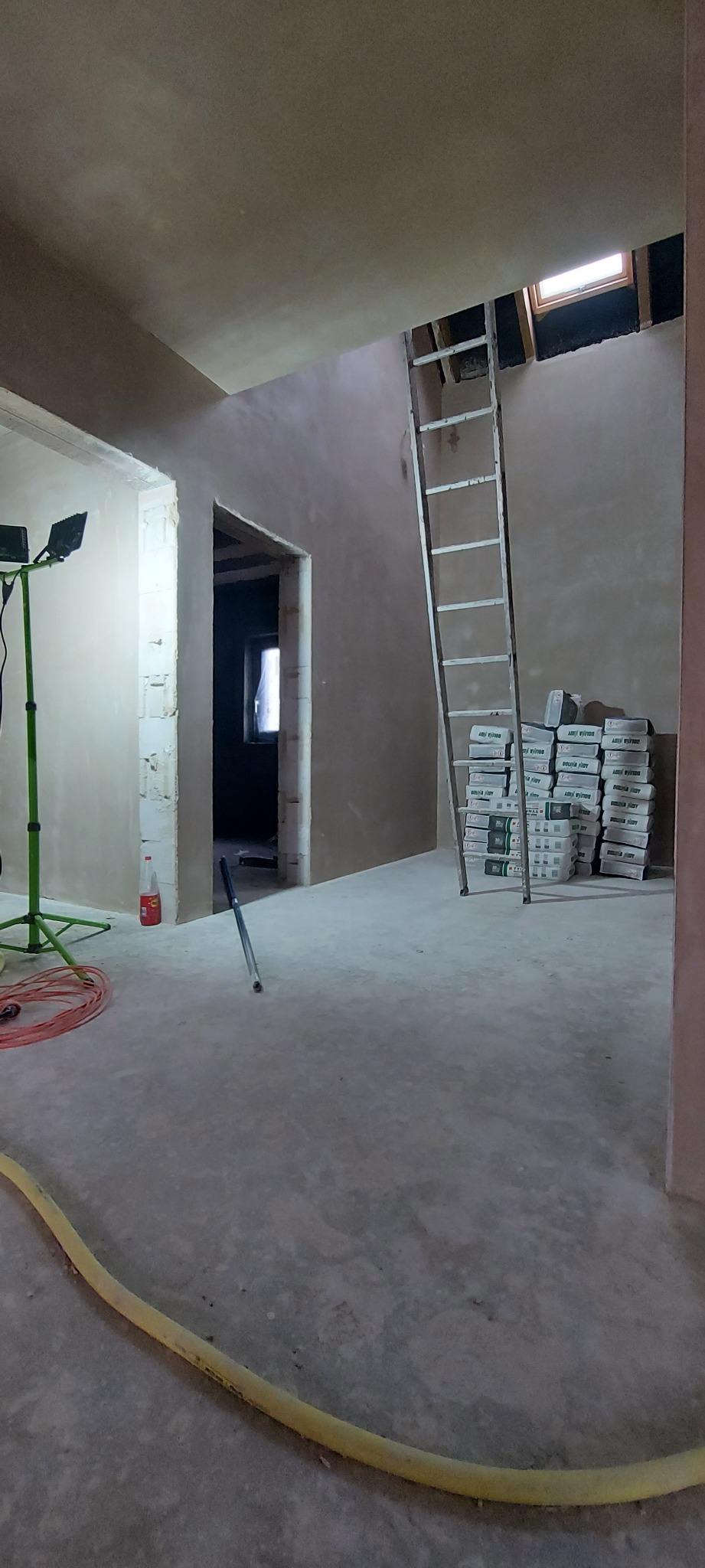 Świeżo położone tynki maszynowe w surowym wnętrzu, widoczna drabina oparta o ścianę i sterta worków z zaprawą, kabel od agregatu tynkarskiego na podłodze.