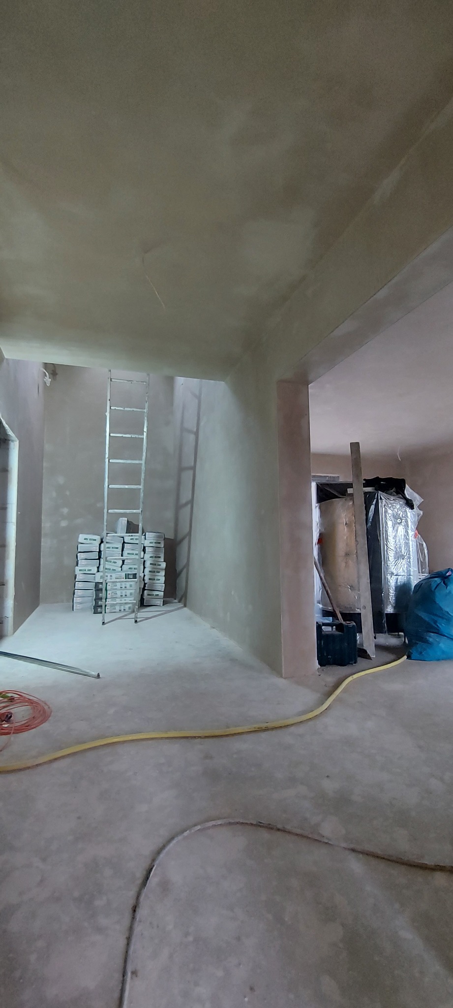 Wnętrze po tynkowaniu maszynowym, widoczny stos worków z zaprawą, drabina oparta o ścianę, żółty wąż i inne narzędzia budowlane.
