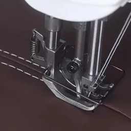 Szczegółowe ujęcie stopki maszyny do szycia, przeszywającej brązową skórę ekologiczną białą nicią, widoczny precyzyjny szew z równymi odstępami.