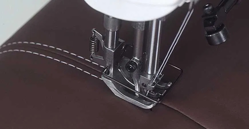 Szczegółowe ujęcie stopki maszyny do szycia, przeszywającej brązową skórę ekologiczną białą nicią, widoczny precyzyjny szew z równymi odstępami.