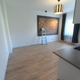 Well done - Jasne panele podłogowe w przestronnym pokoju z drabiną opartą o ceglaną ścianę w ramie. Widoczne lampy i okno z zasłonami. Część blatu kuchennego w kadrze.