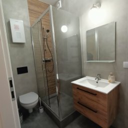 Well done - Nowoczesna łazienka w szarościach z drewnianymi akcentami: kabina prysznicowa, umywalka z szafką, toaleta. Minimalistyczny design z dbałością o detale.