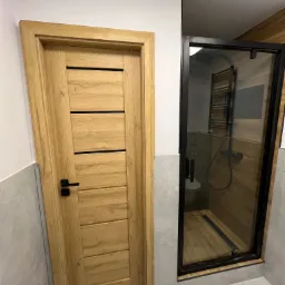 Nowoczesna łazienka z drewnianymi drzwiami z czarną klamką i kabiną prysznicową walk-in z czarnymi profilami, widoczny grzejnik drabinkowy i drewniane wykończenie wewnątrz kabiny.
