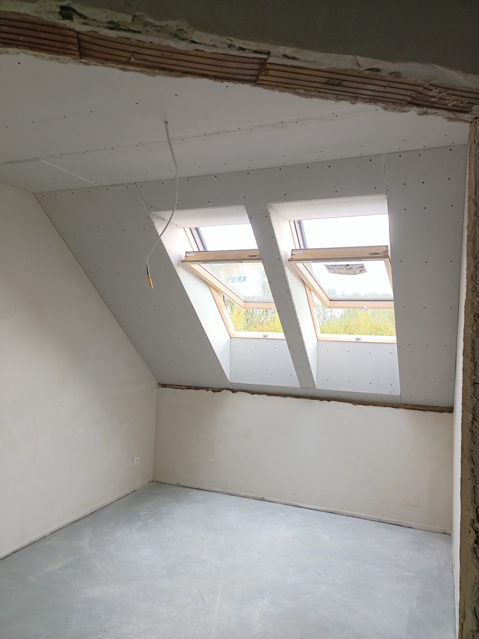 Poddasze z dwoma oknami dachowymi, wykończone płytami karton-gips, widoczne fragmenty konstrukcji drewnianej stropu, surowa wylewka na podłodze, ściany przygotowane do malowania.