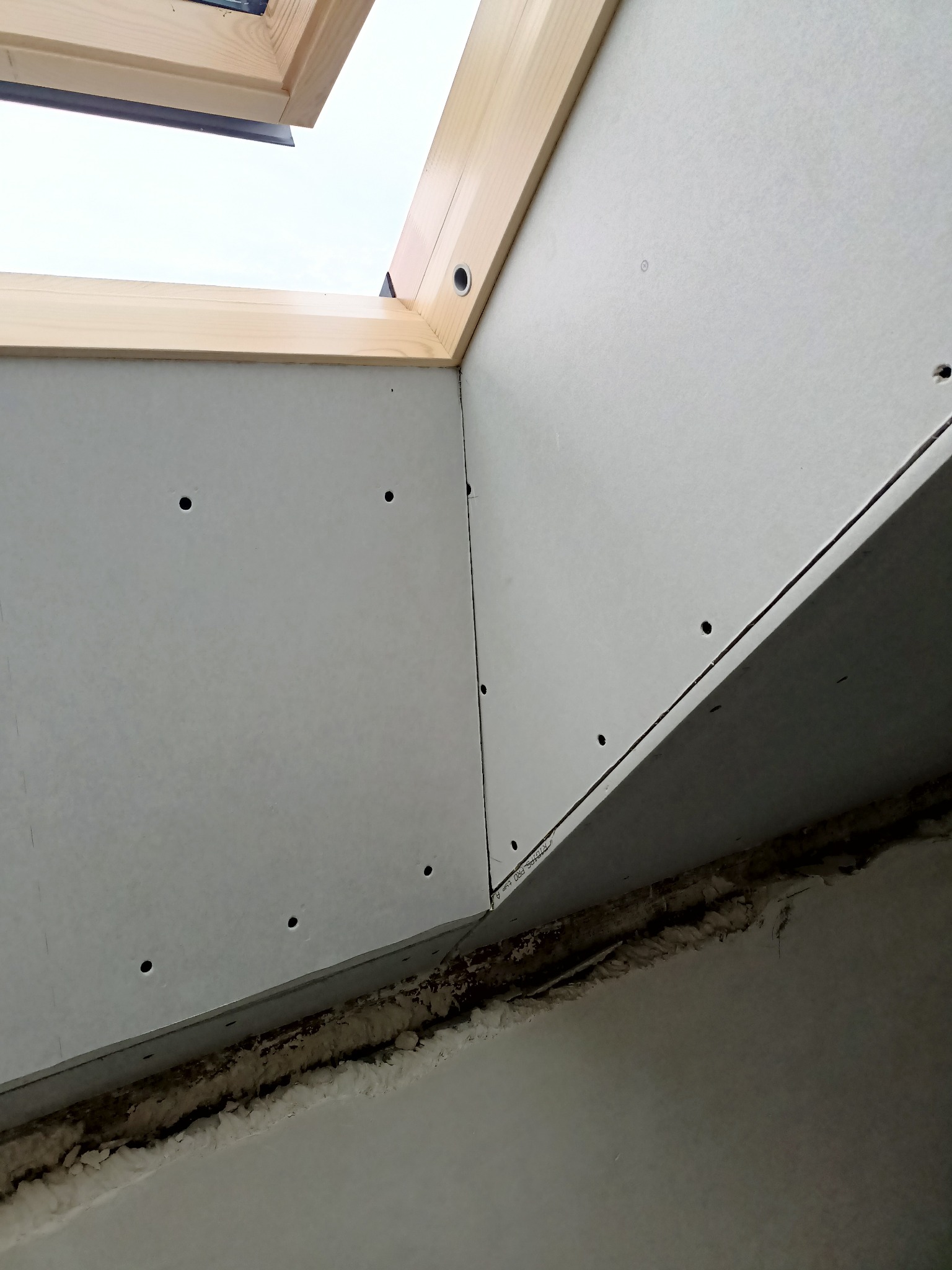 Montaż płyty gipsowo-kartonowej wokół okna dachowego, widoczne otwory po wkrętach i pianka izolacyjna w szczelinie.