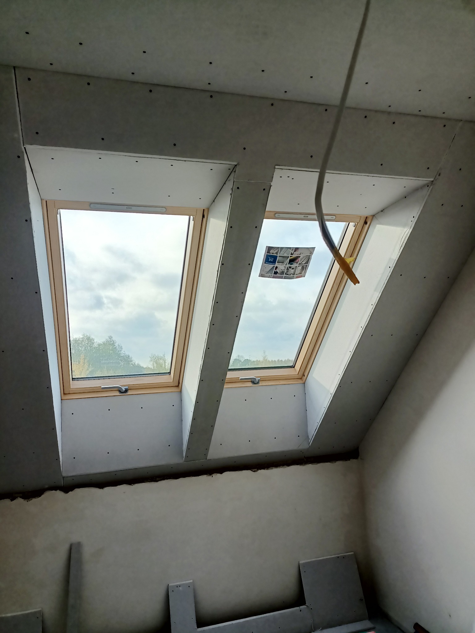 Poddasze z dwoma oknami dachowymi w drewnianych ramach, obudowane płytami kartonowo-gipsowymi, widoczne ślady szpachlowania, kabel zwisający z sufitu.