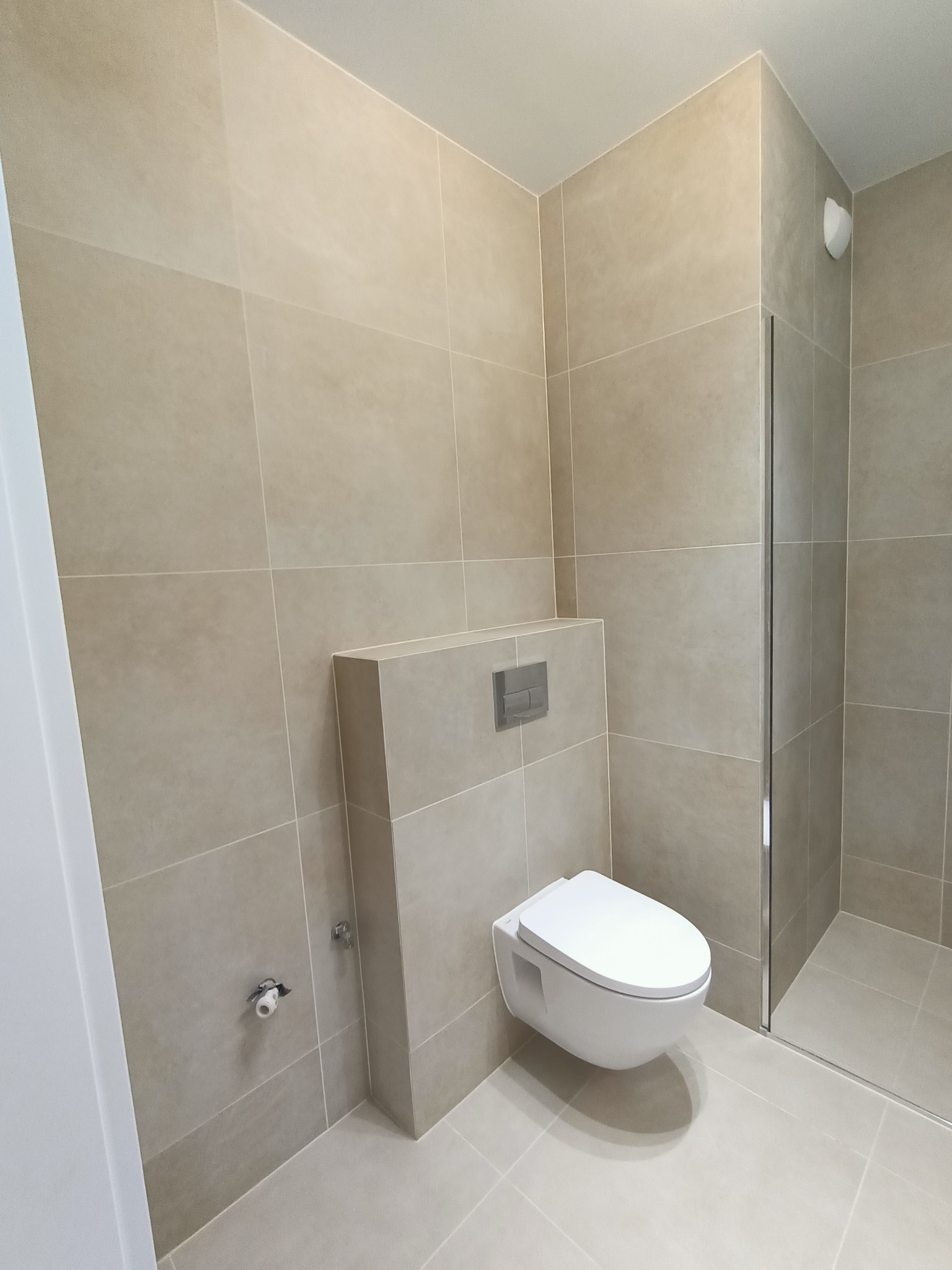 Beżowa łazienka z podwieszaną toaletą i kabiną prysznicową. Minimalistyczny design, płytki w odcieniach beżu, nowoczesna armatura. Widok na fragment kabiny.