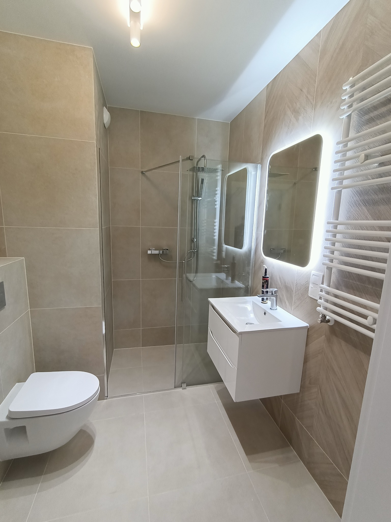 Nowoczesna łazienka w beżach: kabina prysznicowa, podwieszana toaleta, umywalka z szafką i podświetlane lustro. Minimalistyczny design, grzejnik drabinkowy.