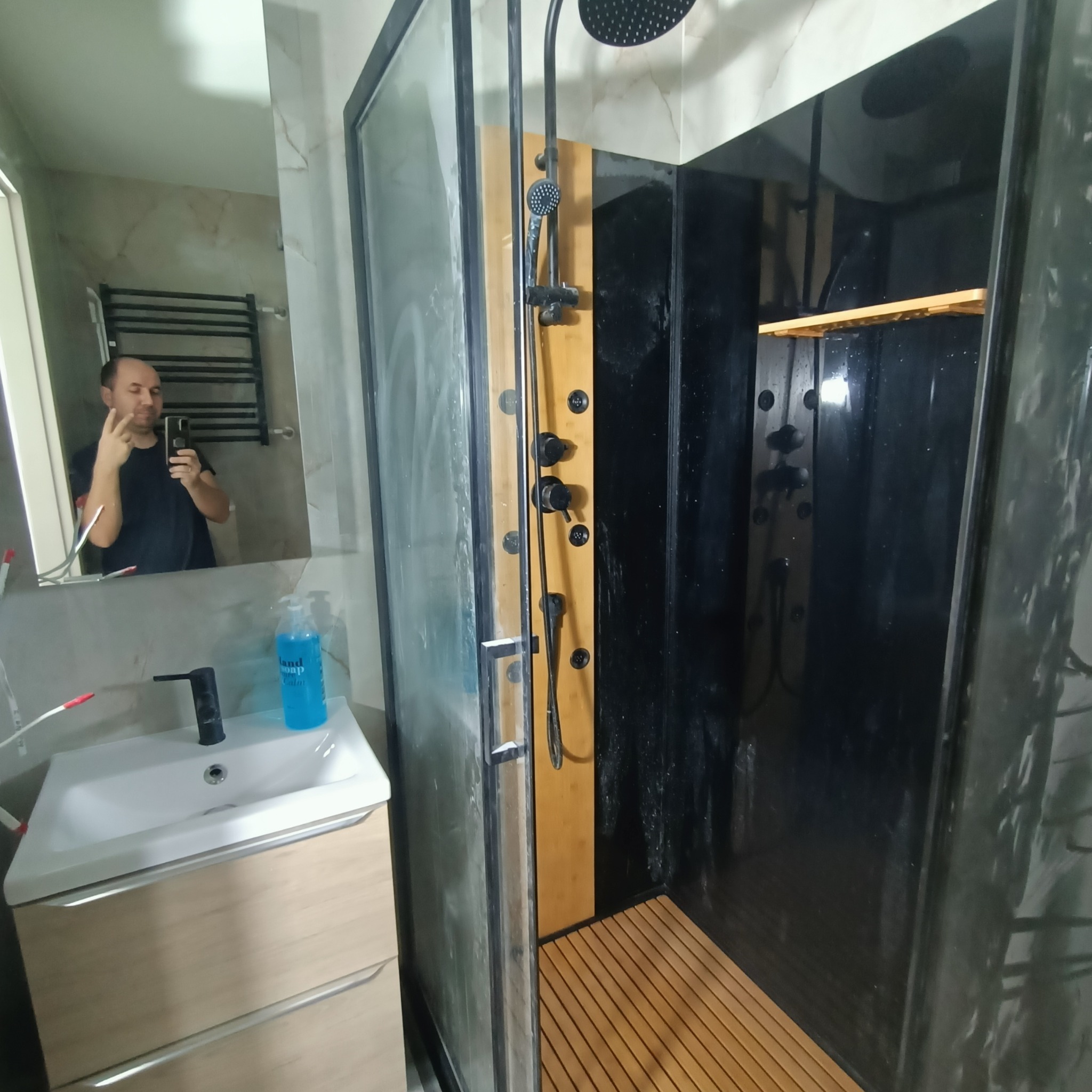 Nowoczesna kabina prysznicowa z czarnymi panelami i drewnianym wykończeniem, widok na umywalkę z szafką i odbicie osoby w lustrze.