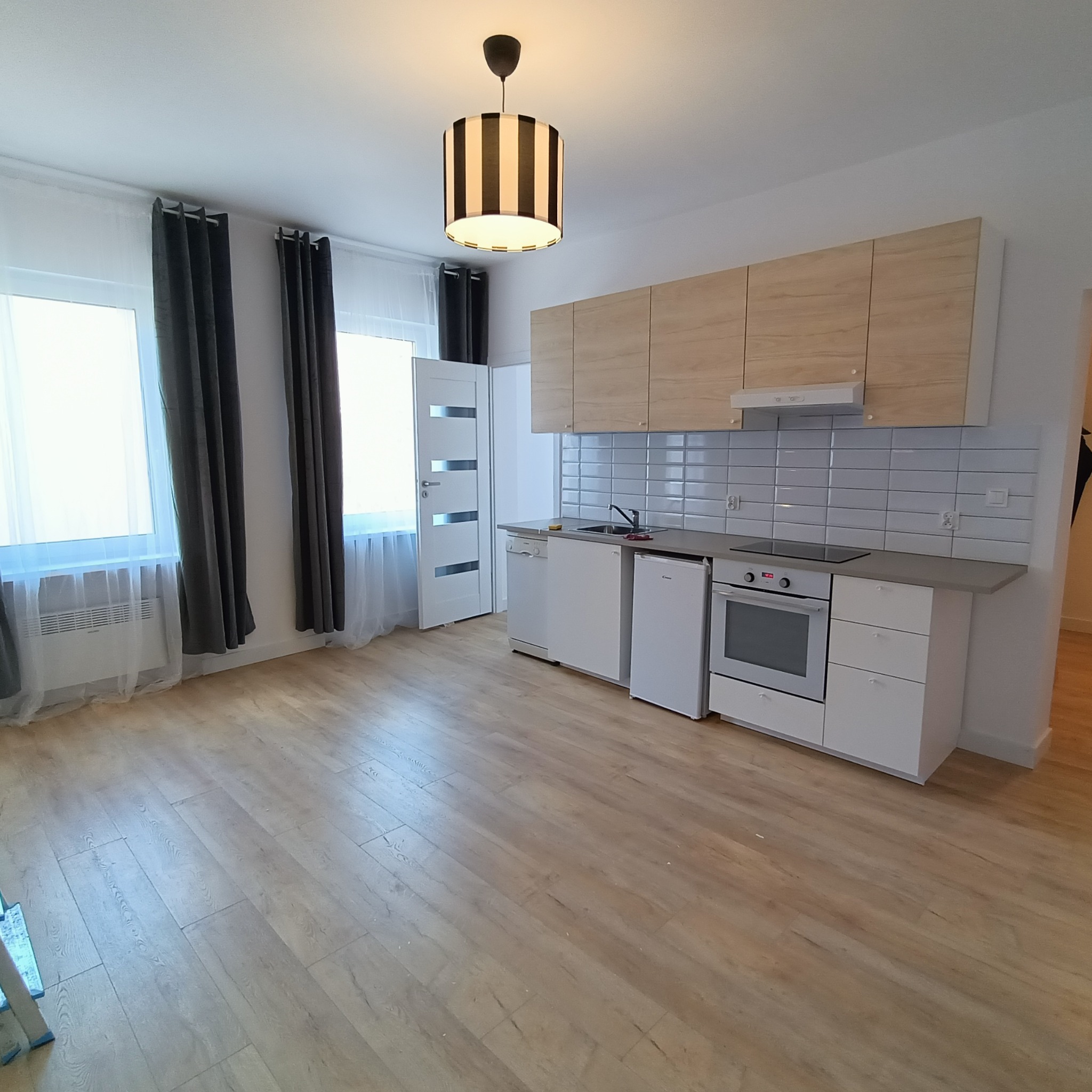 Jasna, nowocześnie urządzona kuchnia z drewnianymi szafkami, białymi sprzętami i panelami podłogowymi. Widoczne okna z ciemnymi zasłonami i lampa w paski.