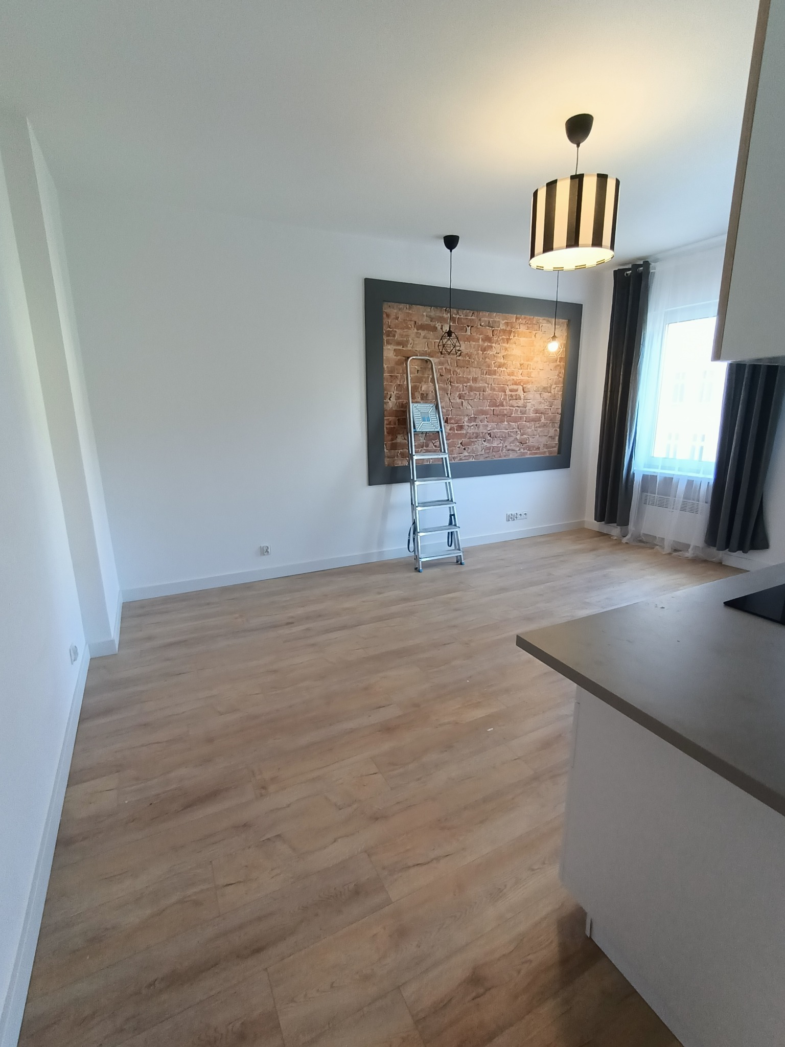 Jasne panele podłogowe w przestronnym pokoju z drabiną opartą o ceglaną ścianę w ramie. Widoczne lampy i okno z zasłonami. Część blatu kuchennego w kadrze.