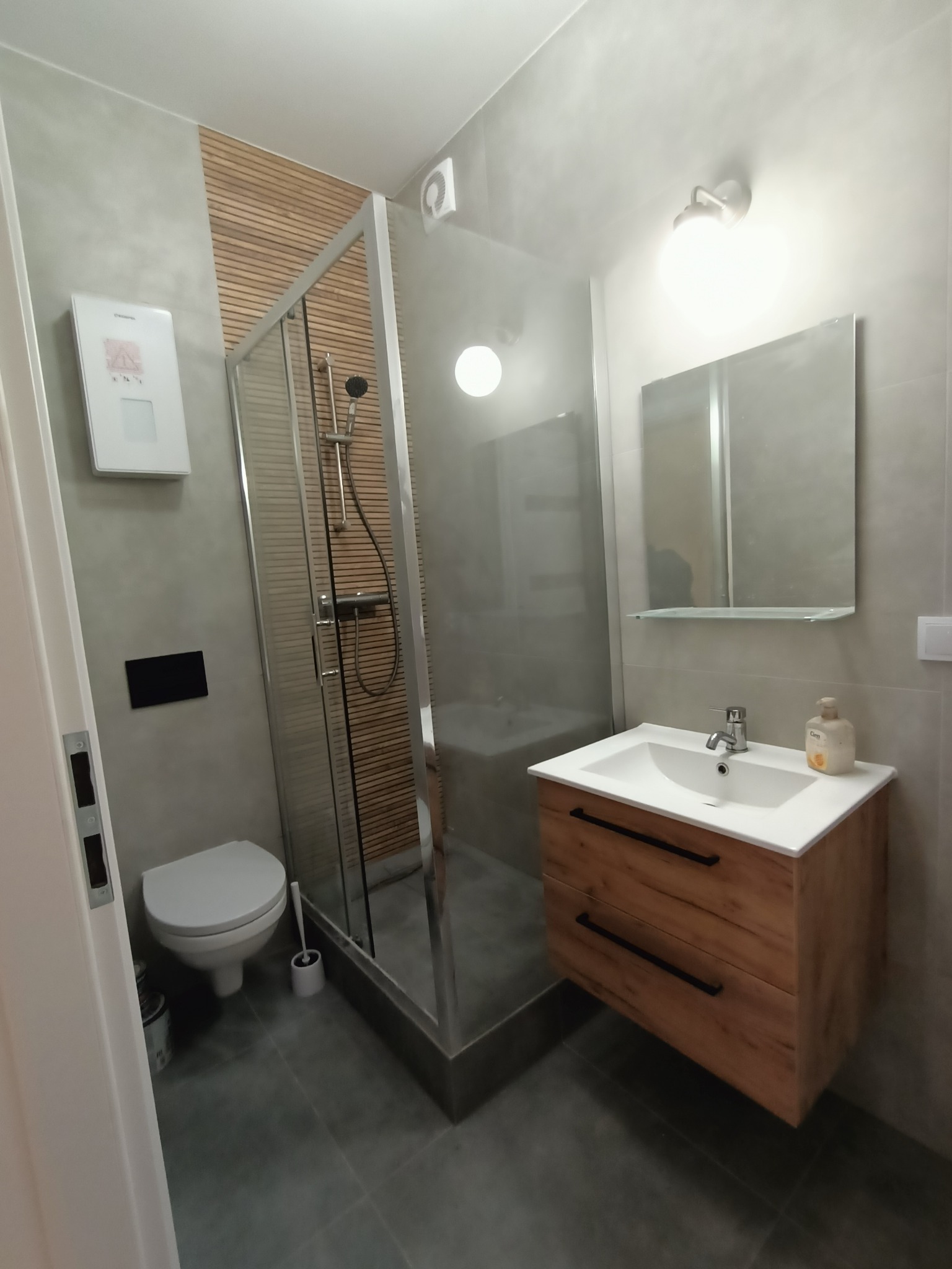 Nowoczesna łazienka w szarościach z drewnianymi akcentami: kabina prysznicowa, umywalka z szafką, toaleta. Minimalistyczny design z dbałością o detale.