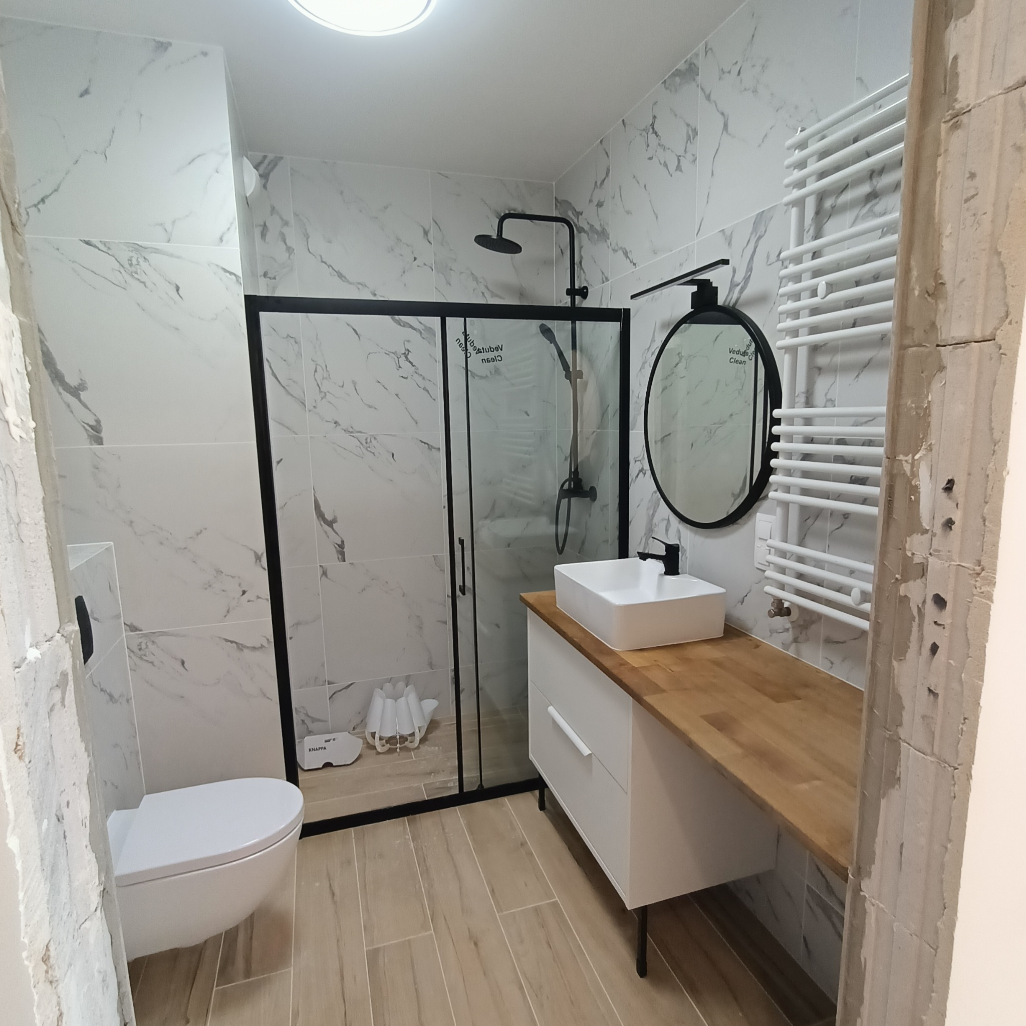 Nowoczesna łazienka z kabiną prysznicową typu walk-in, umywalką nablatową na drewnianym blacie i grzejnikiem drabinkowym. Marmurowe płytki na ścianach.