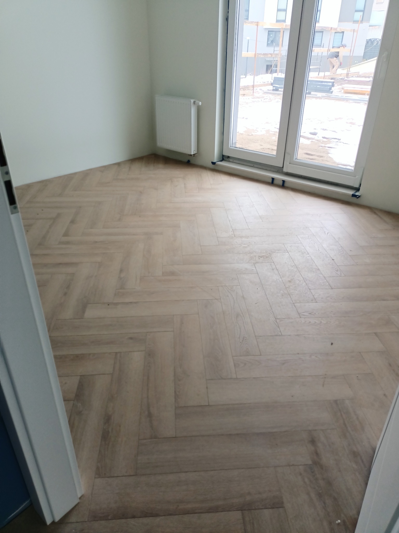 Jasne panele podłogowe ułożone we wzór jodełki francuskiej w nowym, pustym pokoju z grzejnikiem i dużym oknem z widokiem na plac budowy.