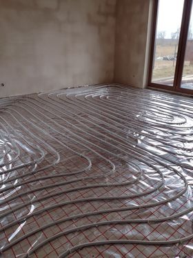 Rozłożone rury ogrzewania podłogowego na folii z siatką w surowym wnętrzu, widok z niskiej perspektywy na układ pętli w dużej przestrzeni.