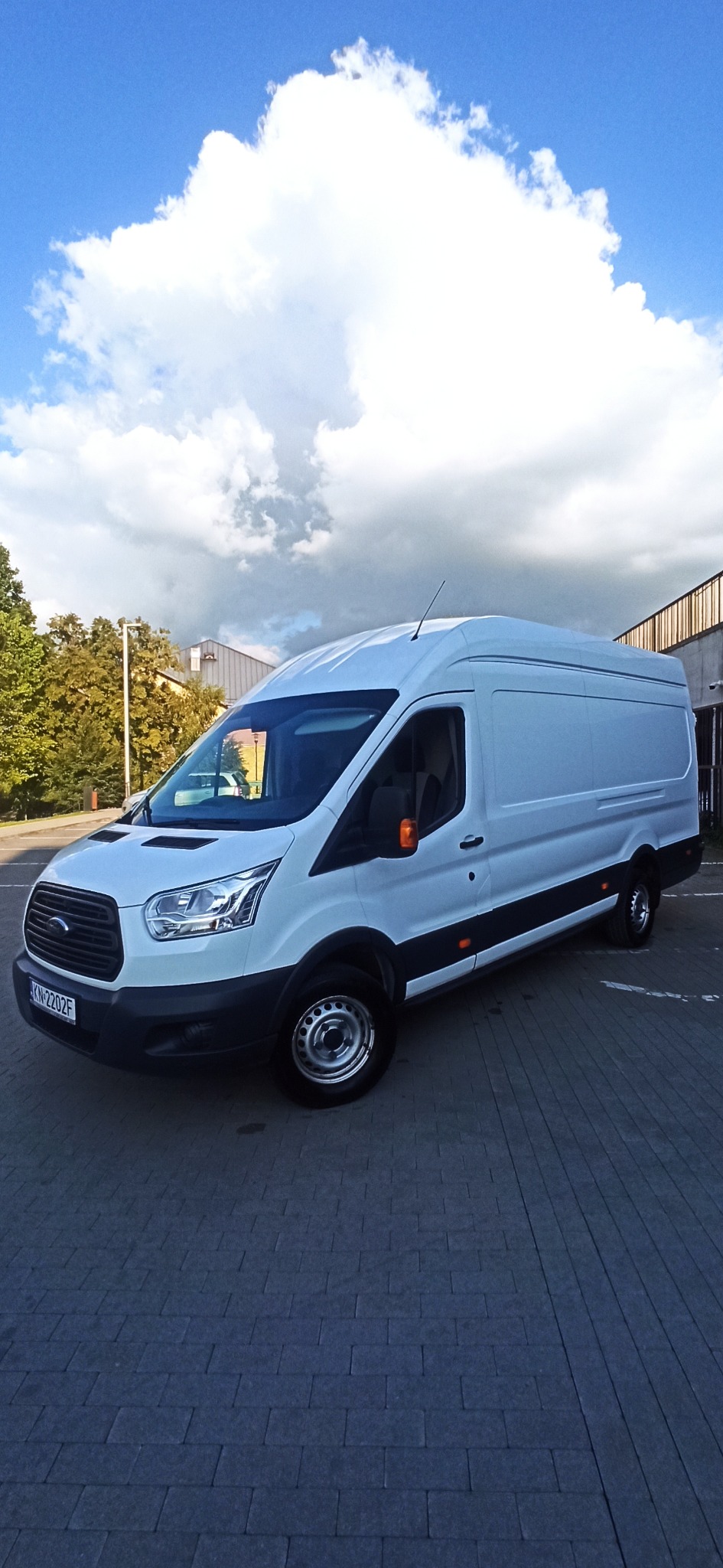 Biały samochód dostawczy Ford Transit zaparkowany na brukowanym placu, z widoczną tablicą rejestracyjną i błękitnym niebem z chmurami w tle.