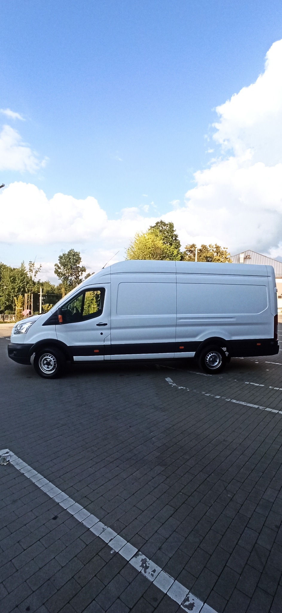 Biały samochód dostawczy zaparkowany na brukowanym parkingu w słoneczny dzień, widok z boku, widoczne drzewa w tle.