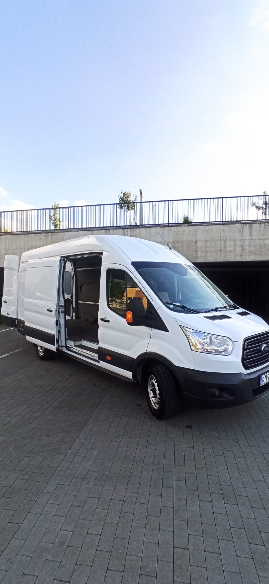 Biały Ford Transit z otwartymi drzwiami bocznymi i tylnymi, zaparkowany na brukowanym placu, widok z boku w słoneczny dzień.