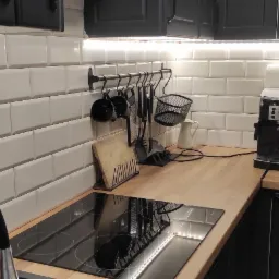 Białe, prostokątne płytki na ścianie w kuchni, podświetlone listwą LED, z czarnymi szafkami i drewnianym blatem, na którym stoi ekspres do kawy.